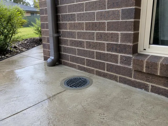 Outdoor drain grate beside brick wall and wet concrete paving in Kellyville